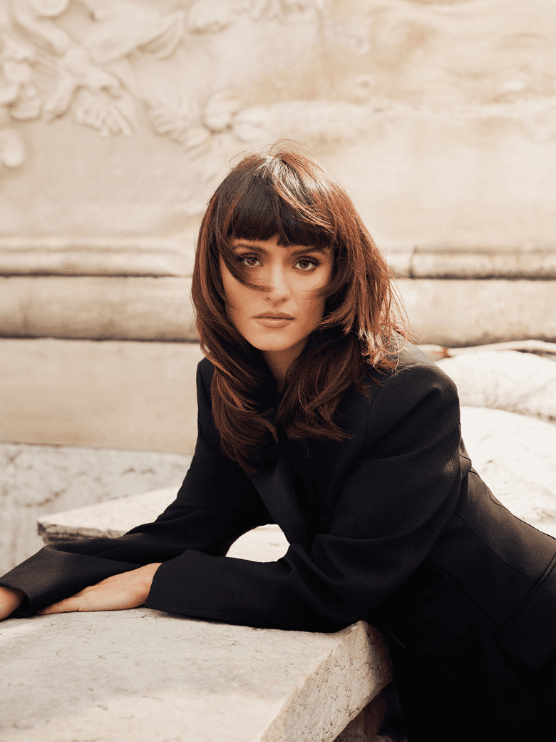 Femme aux cheveux bruns mi-longs avec une frange droite, portant un blazer noir sortant d'un salon Franck Provost après une coloration avec les produits iNOA et Dia color de l'Oréal Professionnel.