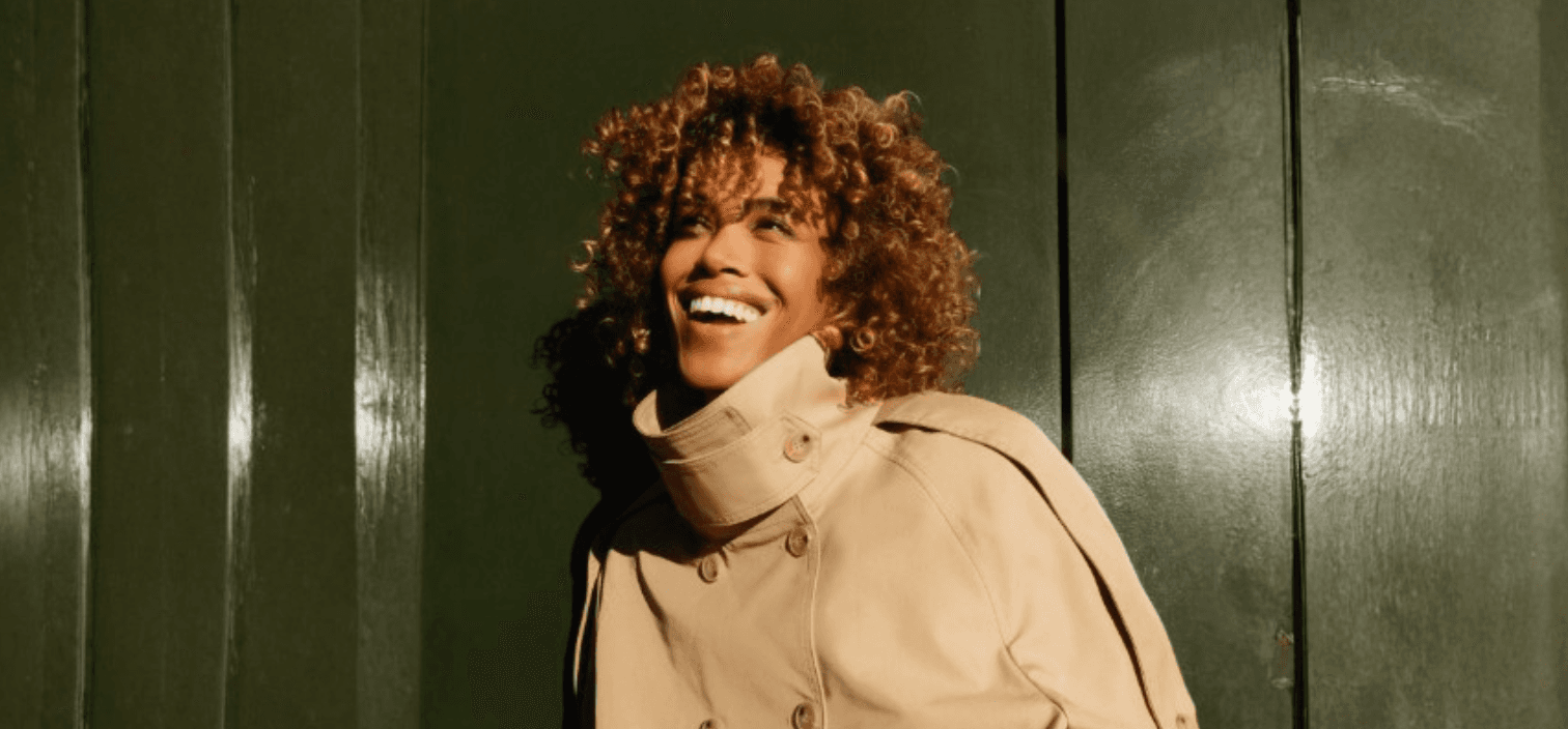 Femme souriante aux cheveux bouclés miel, portant un trench beige, mettant en avant la définition des boucles Franck Provost.